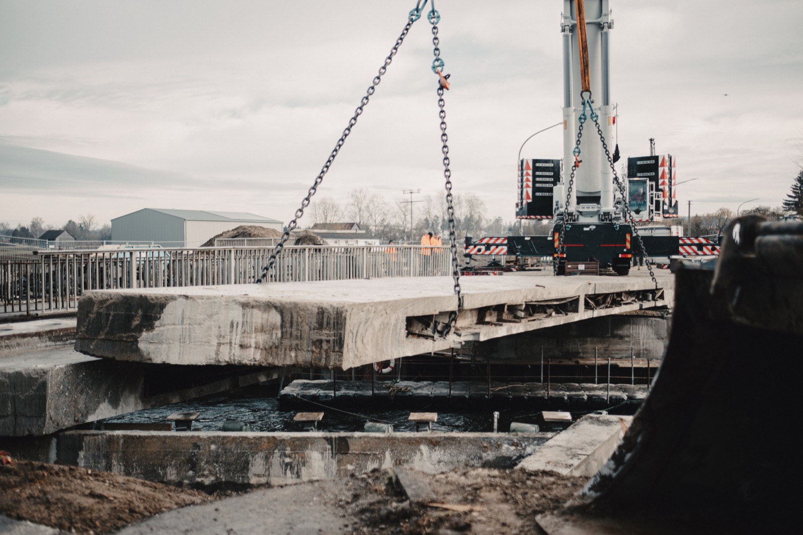 Kran hebt Brückensegment während Bagger am Ufer bereitsteht
